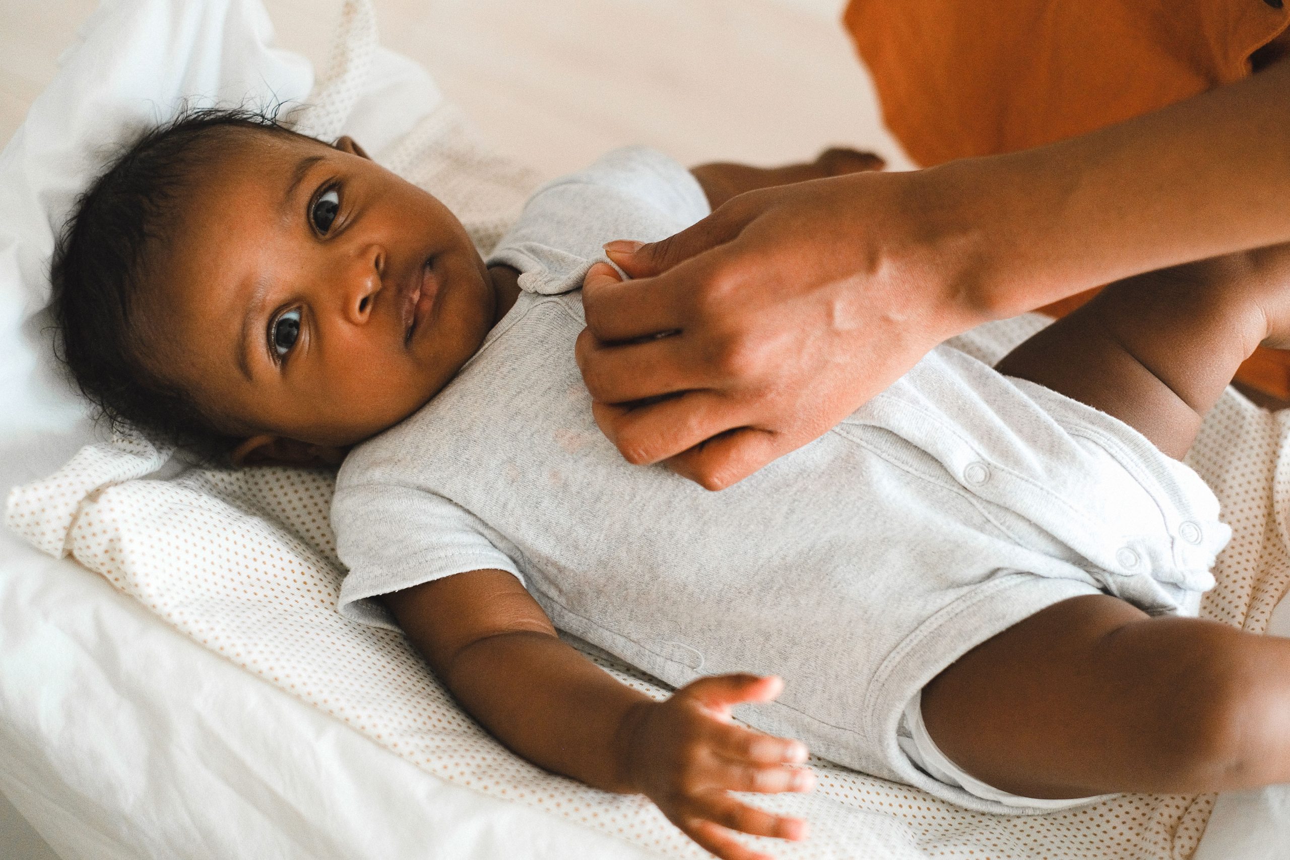 Baby lying down maintaining eye contact with the camera, with an adult's hand resting gently on the baby's chest